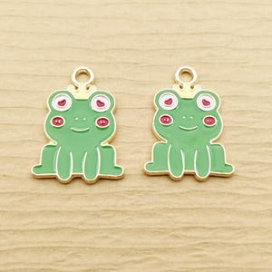 May include: Two identical metal charms shaped like green frogs wearing small gold crowns. Each frog features white eye sockets with red heart-shaped pupils, red circular cheek markings, and a gold-toned metal outline with a top loop for hanging.