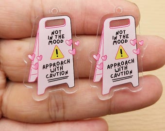 May include: Two clear acrylic charms shaped like caution signs. Each sign is pink with the text "NOT IN THE MOOD", a yellow exclamation point, and "APPROACH WITH CAUTION". Pink hearts decorate the sides.