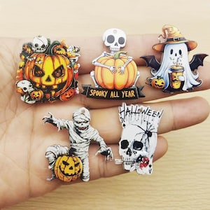 May include: Five flat Halloween-themed charms featuring a jack-o'-lantern with skulls, a skeleton holding a pumpkin with the text SPOOKY ALL YEAR, a ghost in a witch hat holding a drink, a mummy with a pumpkin, and a skull on a playing card.