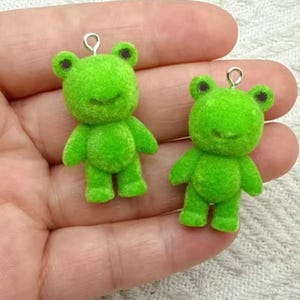 May include: Two bright green frog-shaped charms with a fuzzy texture. Each charm has a silver-colored loop at the top for hanging. The frogs have small black eyes and smiling faces. These charms are suitable for jewelry making or craft projects.