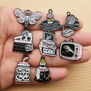 May include: A collection of black, pink, and white enamel charms with a spooky theme. Designs include a moth, records with a rose, a gramophone, a film clapper, a typewriter with the word "HELP", a television with the word "COMING", a jar of skulls, and a potion bottle.