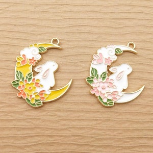May include: Two gold-tone metal charms shaped like a crescent moon with a white rabbit sitting on it. One charm has yellow flowers and the other has pink flowers.