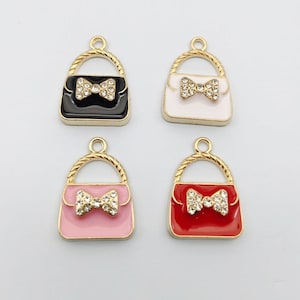 May include: Four handbag charms with gold-tone metal frames and twisted handles. Each charm features a central bow embellished with clear rhinestones. The bags are finished in solid black, white, light pink, and red enamel colours respectively.