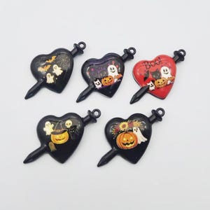 May include: Five heart-shaped charms with a pointed tip and a top loop. Four are black and one is red. The designs feature Halloween motifs including ghosts, pumpkins, bats, a spiderweb, a skull, a black cat, and the text Boo on the red charm.