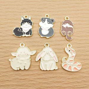 May include: Six gold-tone metal charms featuring different animal designs. The charms include a black and white cat, a black and white hamster, a brown otter, a white lamb, a white rabbit, and a pink and white snake.