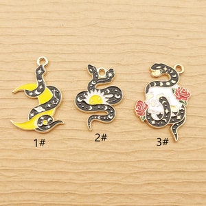 May include: Three gold-toned metal snake charms with black and white enamel designs. The first charm features a snake wrapped around a crescent moon with stars. The second charm features a snake wrapped around a sun with stars. The third charm features a snake wrapped around a skull with roses.