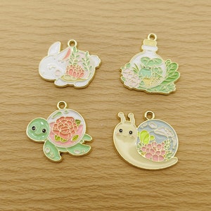 May include: Four gold-tone metal charms featuring a white rabbit, a green turtle, a yellow snail, and a green frog inside a glass bottle. Each charm has a pink flower and green leaves inside a clear dome.