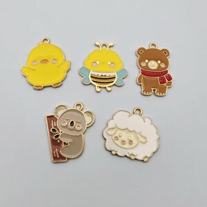 May include: Five metal enamel charms featuring cartoon animals and insects. The set includes a yellow chick, a yellow and blue bee, a brown bear wearing a red scarf, a grey koala holding a brown branch, and a white fluffy sheep with gold outlines.