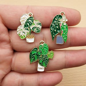 May include: Three gold-tone metal charms featuring different potted plants. The charms are enamel painted in shades of green, white, and blue. The charms have a loop at the top for attaching to a necklace or bracelet. The charms feature the text "PLANTS".