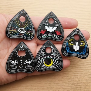 May include: Five black triangular planchette charms with different designs.  The designs include a rainbow eye, a bat, a cat, a crescent moon, and a cat reading a book.  Each planchette has the words "Hello", "Goodbye", "Yes", and "No" written on it.
