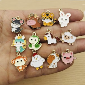 May include: A collection of twelve gold-toned metal charms featuring colorful enamel designs of various animals, including a mouse, ox, tiger, rabbit, dragon, snake, horse, sheep, monkey, rooster, dog, and pig, each with a small loop for attachment.