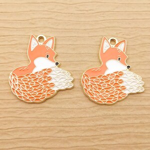 May include: Two gold-colored metal charms shaped like foxes. The foxes are orange with white accents and have detailed patterns on their fur.