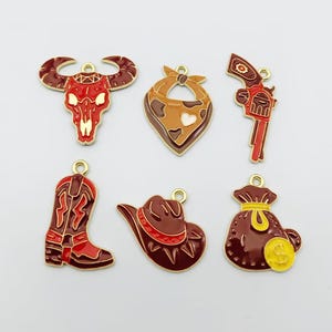 Könnte beinhalten: Ein Set aus sechs Metallanhängern, bestehend aus einem roten Stierschädel, einem braun gemusterten Bandana, einem rot-goldenen Revolver, einem roten Cowboystiefel, einem braunen Cowboyhut und einem braunen Geldsack mit einem gelben Dollarzeichen.