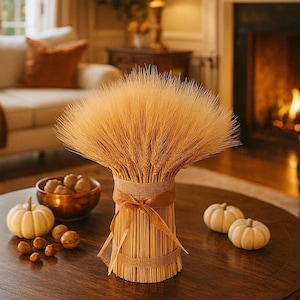 May include: A cozy living room scene with a round wooden coffee table. A bundle of wheat stalks tied with burlap and ribbon sits on the table, along with small white pumpkins and a bowl of nuts. A fireplace and sofa are in the background.