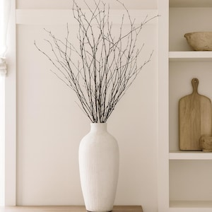 May include: A tall, white ceramic vase filled with dark brown branches. The vase sits on a wooden table with three drawers. The background is a neutral-toned wall with a built-in shelf holding decorative items.