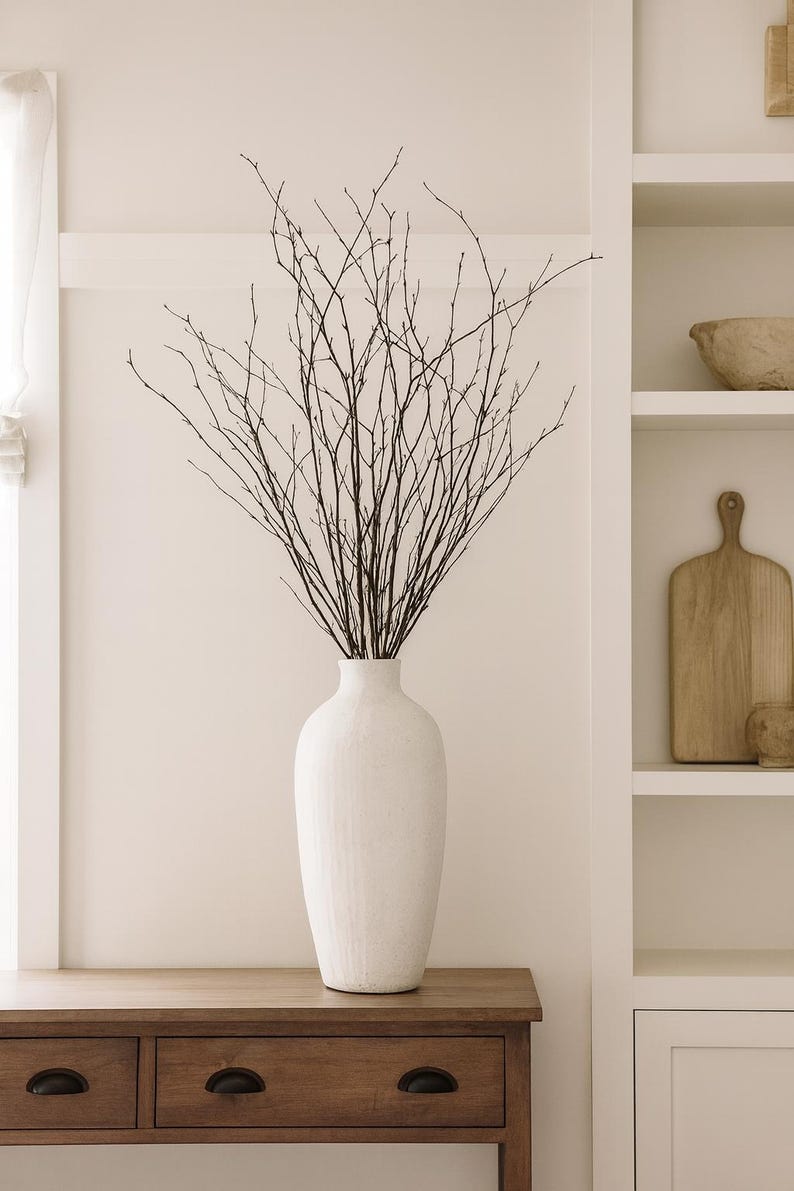 K&ouml;nnte beinhalten: Eine hohe, wei&szlig;e Keramikvase gef&uuml;llt mit dunkelbraunen Zweigen. Die Vase steht auf einem Holztisch mit Schubladen. Der Hintergrund ist eine neutral get&ouml;nte Wand mit einem Einbauregal.