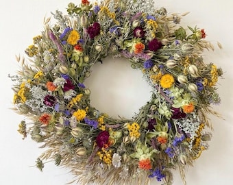 May include: A circular wreath composed of dried flowers and grasses in a vibrant palette of yellow, deep purple, orange, and muted green. The arrangement features various textures including small yellow blossoms, purple petals, and dried oat stalks.