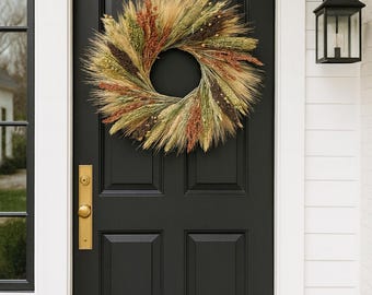 May include: A decorative wreath made of dried wheat, grasses, and seed heads hangs on a black front door. The wreath features a circular design with natural colors of brown, green, and tan. A gold-colored door handle and a black lantern are also visible.