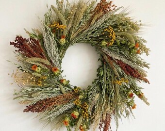 May include: A circular wreath composed of dried grasses, wheat stalks, and various grains in shades of sage green, muted gold, and deep rust brown. The arrangement features textured seed heads, thin stems, and small clusters of yellow and orange flowers.