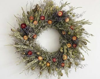 May include: A circular wreath composed of dried botanical elements including green grasses, thin stems, and small seed pods. The arrangement is accented with small, vibrant dried flowers in shades of deep red, burnt orange, and muted yellow throughout.