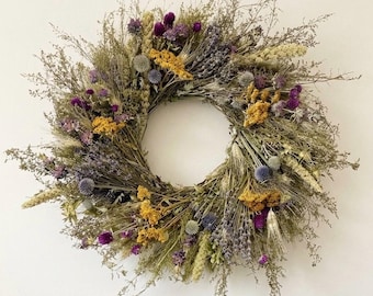 May include: A circular wreath composed of dried botanical elements including purple globe amaranth, yellow yarrow, blue globe thistle, and sprigs of lavender. The arrangement features a variety of dried grasses and stems in shades of green and brown.