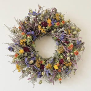 Op de afbeelding: Een ronde krans samengesteld uit gedroogde bloemen en bladeren in een levendige mix van kleuren. Het arrangement bevat paarse, gele, oranje en donkerrode bloesems afgewisseld met zilvergroene gedroogde stengels, zaaddozen en textuurrijk groen.