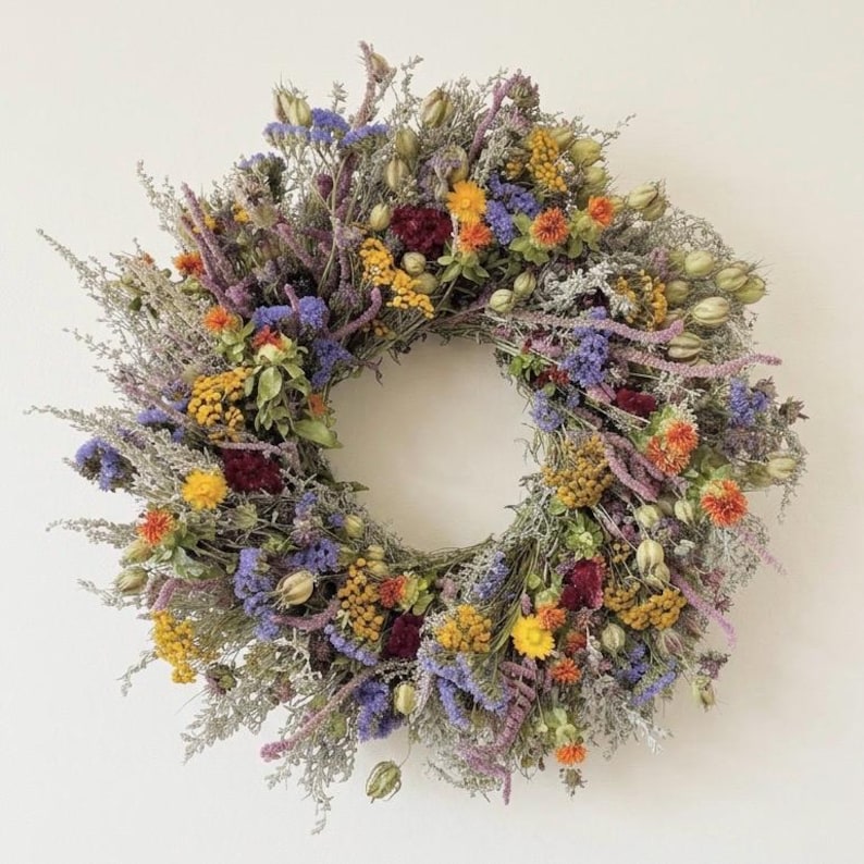 Op de afbeelding: Een ronde krans samengesteld uit gedroogde bloemen en bladeren in een levendige mix van kleuren. Het arrangement bevat paarse, gele, oranje en donkerrode bloesems afgewisseld met zilvergroene gedroogde stengels, zaaddozen en textuurrijk groen.