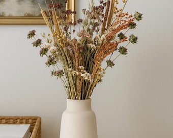 May include: A cream-colored vase holds a bouquet of dried flowers and grasses. The arrangement includes various textures and colors, such as beige, brown, and green. The vase sits on a white surface, with a framed landscape painting in the background.