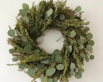 Pode incluir: Uma guirlanda circular construída a partir de um arranjo denso de folhagem verde seca e folhas de eucalipto. O design apresenta uma mistura de caules de plantas plumosos e texturizados com folhas de eucalipto lisas e arredondadas em tons.