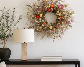 May include: A colourful dried flower wreath hangs on a white wall. A black console table holds a lamp with a beige shade, books, and a decorative bowl. A potted plant and a sofa are also visible. The scene is well-lit.