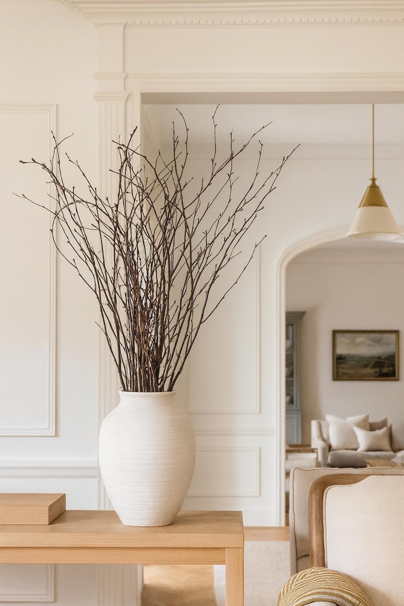 K&ouml;nnte beinhalten: Eine gro&szlig;e, cremewei&szlig;e Keramikvase gef&uuml;llt mit zahlreichen braunen Zweigen. Die Vase steht auf einem hellen Holztisch, mit einem Blick auf ein Wohnzimmer im Hintergrund. Der Raum hat wei&szlig;e W&auml;nde und eine neutrale Farbpalette.