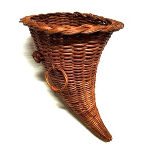 May include: A brown wicker cornucopia with a ring handle. The cornucopia is shaped like a horn and is made of woven wicker.