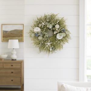 May include: A decorative wreath featuring dried grasses, starfish, and sand dollars. The wreath is adorned with blue and white accents, creating a coastal theme. It is displayed on a white shiplap wall, adding a touch of natural elegance to the space.