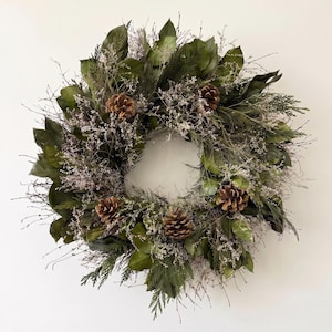 May include: A circular wreath constructed from dark green magnolia leaves, thin brown twigs, and wispy dried white filler flowers. Four brown pinecones are evenly spaced throughout the foliage, creating a textured, natural, and round arrangement.