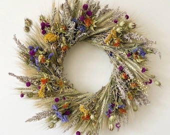 May include: A circular wreath composed of dried wheat, lavender, and colorful wildflowers. The wreath features a variety of colors, including purple, orange, yellow, and blue, creating a natural and rustic aesthetic. Perfect for home decor.