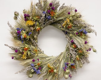 Könnte beinhalten: Ein kreisförmiger Kranz aus getrockneten Weizenhalmen, verziert mit einem Arrangement aus getrockneten Blumen in leuchtenden Violett-, Orange-, Gelb- und Blautönen, durchsetzt mit silberfarbenem Blattwerk und kleinen Samenkapseln.