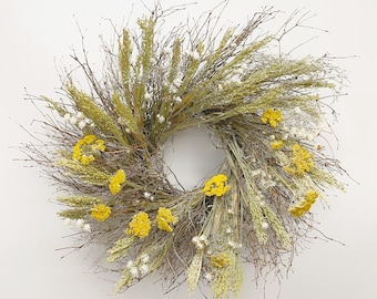 Könnte beinhalten: Ein Trockenblumenkranz aus Zweigen, getrockneten Gräsern und gelben Blüten. Der Kranz ist rund und hängt an einer weißen Wand.