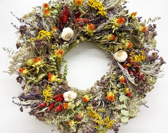 Pode incluir: Uma coroa de flores secas com pimentas vermelhas, flores amarelas e dentes de alho brancos. A coroa é feita de várias ervas e flores secas.