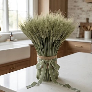 May include: A large, dense bouquet of dried green wheat stalks arranged in a vertical bundle. The base of the wheat is wrapped in a wide band of natural tan burlap, which is tied with a soft, sage green velvet ribbon featuring a decorative bow.