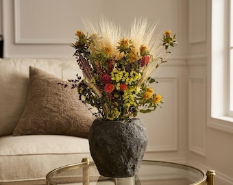 May include: A dark grey, textured, stone-like vase containing a dried floral arrangement. The bouquet features tall beige wheat stalks, bright yellow safflowers, small red globe amaranth blossoms, and clusters of tiny yellow and dark purple flowers.
