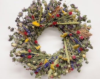 May include: A circular wreath composed of various dried flowers and grasses. The arrangement features a mix of purple, yellow, and red blossoms interspersed with green stems, seed pods, and textured foliage, creating a dense, multi-colored display.