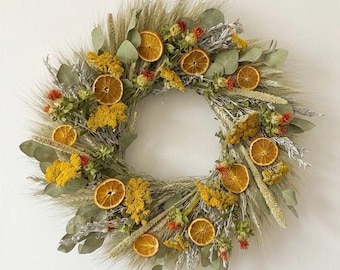 May include: A circular wreath constructed from dried wheat stalks, green eucalyptus leaves, and clusters of small yellow flowers. The arrangement is accented with several dried orange slices and small red floral buds distributed throughout the foliage.