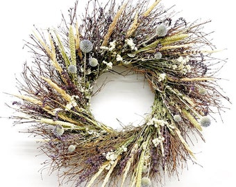 May include: A circular wreath composed of dried branches, grasses, and flowers. The wreath features a variety of textures and colors, including brown, beige, purple, and white. The arrangement is suitable for seasonal home decor.