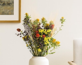 May include: A white ceramic vase filled with a vibrant arrangement of dried flowers. The bouquet includes yellow, orange, and purple blooms, with feathery grasses. A small wooden container sits nearby. A framed landscape painting hangs in the background.