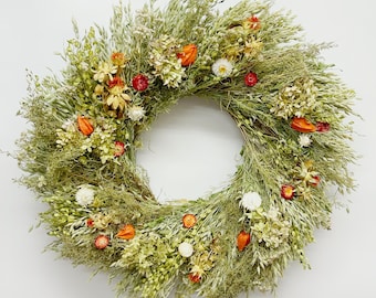 Pode incluir: Uma coroa circular composta por ervas secas, pequenas flores brancas e detalhes em laranja e amarelo. A coroa está sobre um fundo branco. Os materiais naturais criam uma estética rústica e orgânica, adequada para decoração de casa.