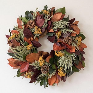 May include: A circular wreath composed of deep green, rust, and brown magnolia leaves interspersed with light green oat grass stalks, small clusters of bright yellow flowers, and several brown pinecones arranged in a dense, layered pattern.
