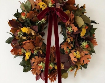 May include: A circular wreath constructed from dried autumn leaves in shades of brown, orange, and deep red, accented with small yellow flower clusters and a long, dark burgundy velvet ribbon tied in a bow at the top center.