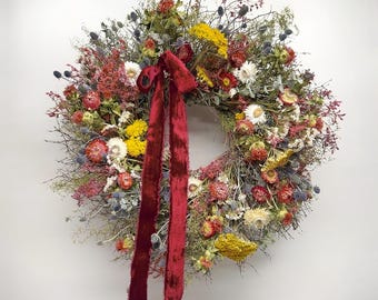 Può includere: Una vivace ghirlanda circolare composta da fiori secchi di vari colori, tra cui rosso, giallo, bianco e blu. Un lungo nastro di velluto rosso scuro è legato alla parte superiore della ghirlanda. La ghirlanda è un oggetto decorativo.