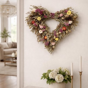 May include: A heart-shaped wreath made of dried flowers and grasses hangs on a white wall. The wreath features a variety of colors, including pink, yellow, and red. Below the wreath is a white vase with white flowers and two gold candle holders.