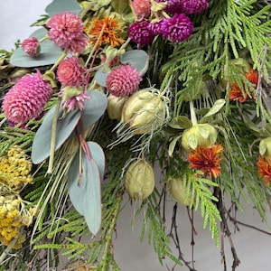 K&ouml;nnte beinhalten: Nahaufnahme eines getrockneten Blumenarrangements mit rosa Kugelamarant, lila Gomphrena, orangefarbenen Bl&uuml;ten und gr&uuml;nem Laub. Das Arrangement umfasst Eukalyptusbl&auml;tter und Samenkapseln und erzeugt eine nat&uuml;rliche, strukturierte &Auml;sthetik.